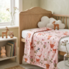 Couverture pour bébé rose poudré à motifs d'animaux de la forêt (faons, hiboux) drapée sur un berceau en bois clair dans une chambre d'enfant d'ambiance sereine et naturelle.