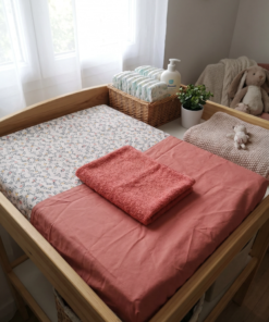 Housse de matelas à langer bicolore à motif floral et tissu terracotta, surmontée de sa serviette en éponge assortie sur une table à langer en bois.